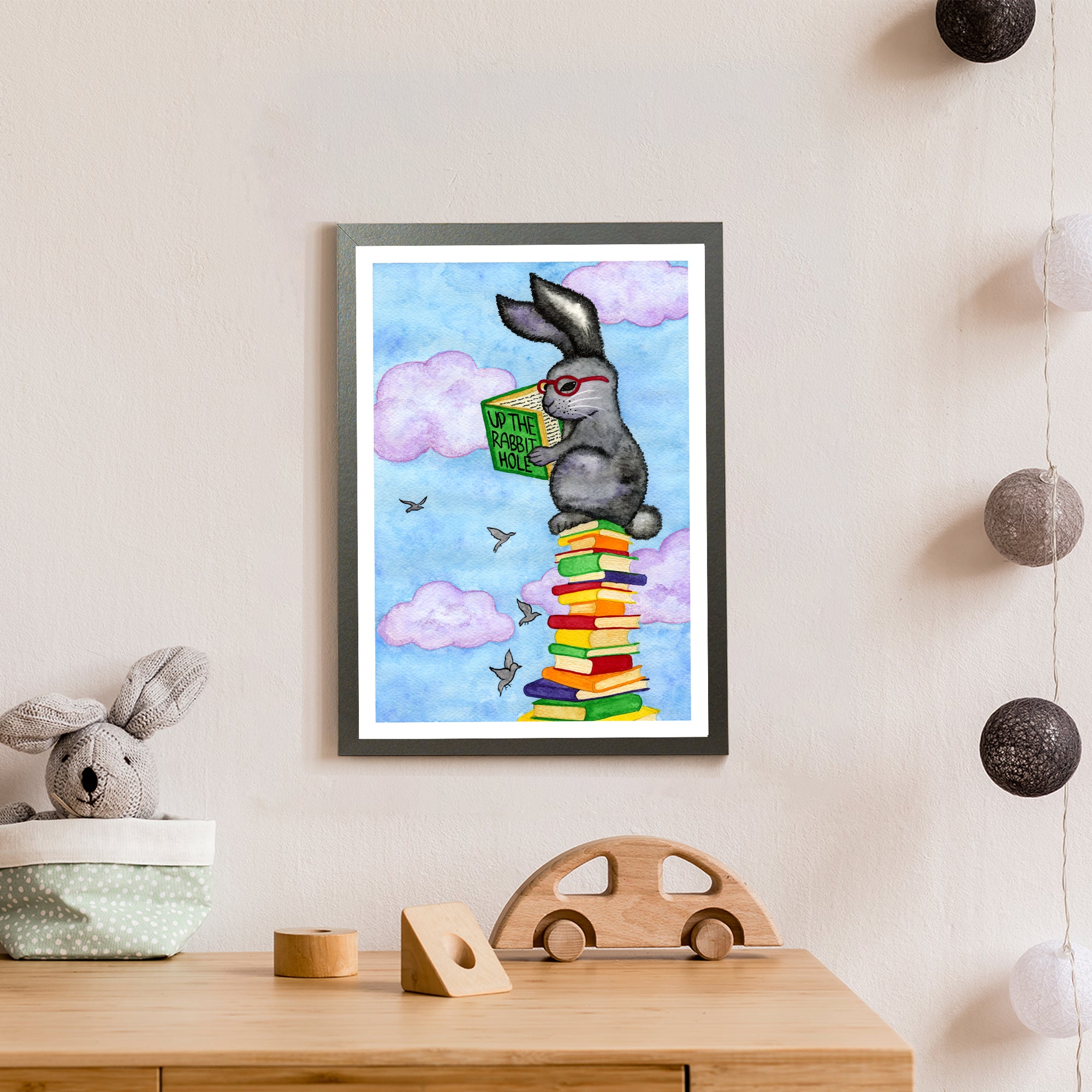 A grey bunny with red glasses reads a book called 'Up the rabbit hole' while sitting on a pile of books so high that he's in the clouds among the birds (print only)