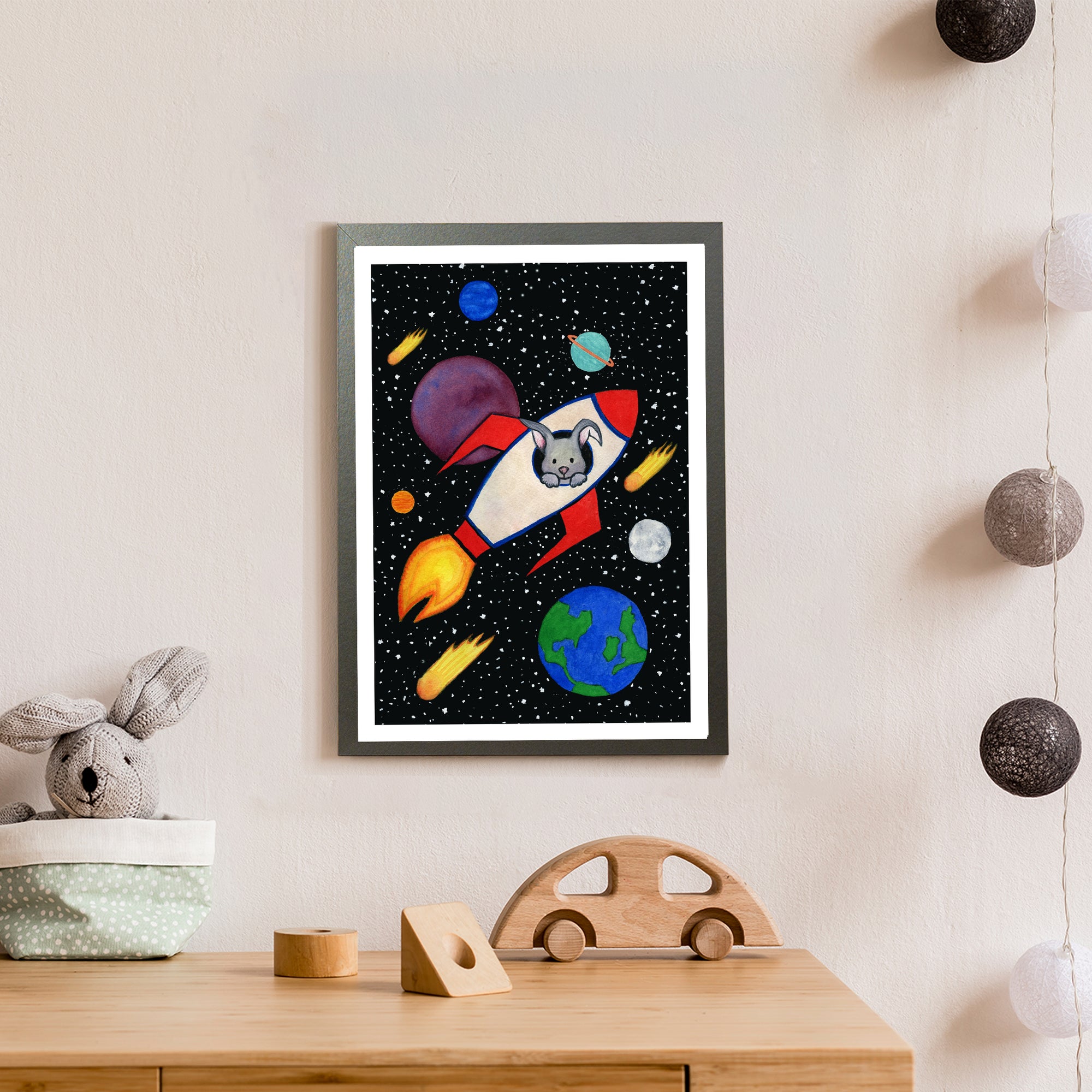 A grey bunny looks out of the window of a red, white and blue rocket, as it flies through space amongst colourful planets, stars and comets (print only)